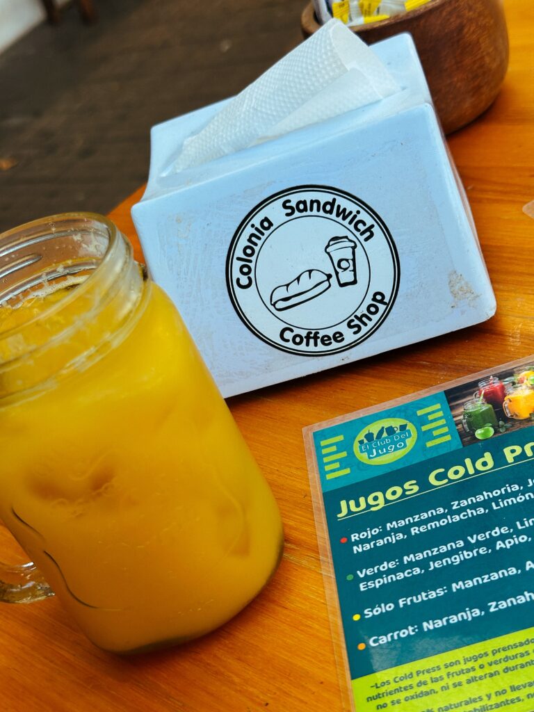 Blue napkin dispenser with a circular 'Colonia Sandwich Coffee Shop' logo, beside a jar of orange juice on a wooden table.