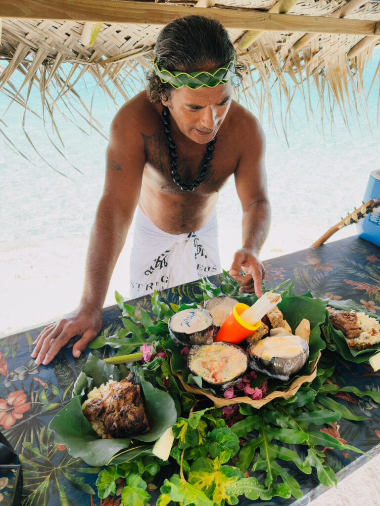 Man at a beach hut arranging a tropical feast on a colorful table, with coconuts, leaves, and tropical fruits nearby.