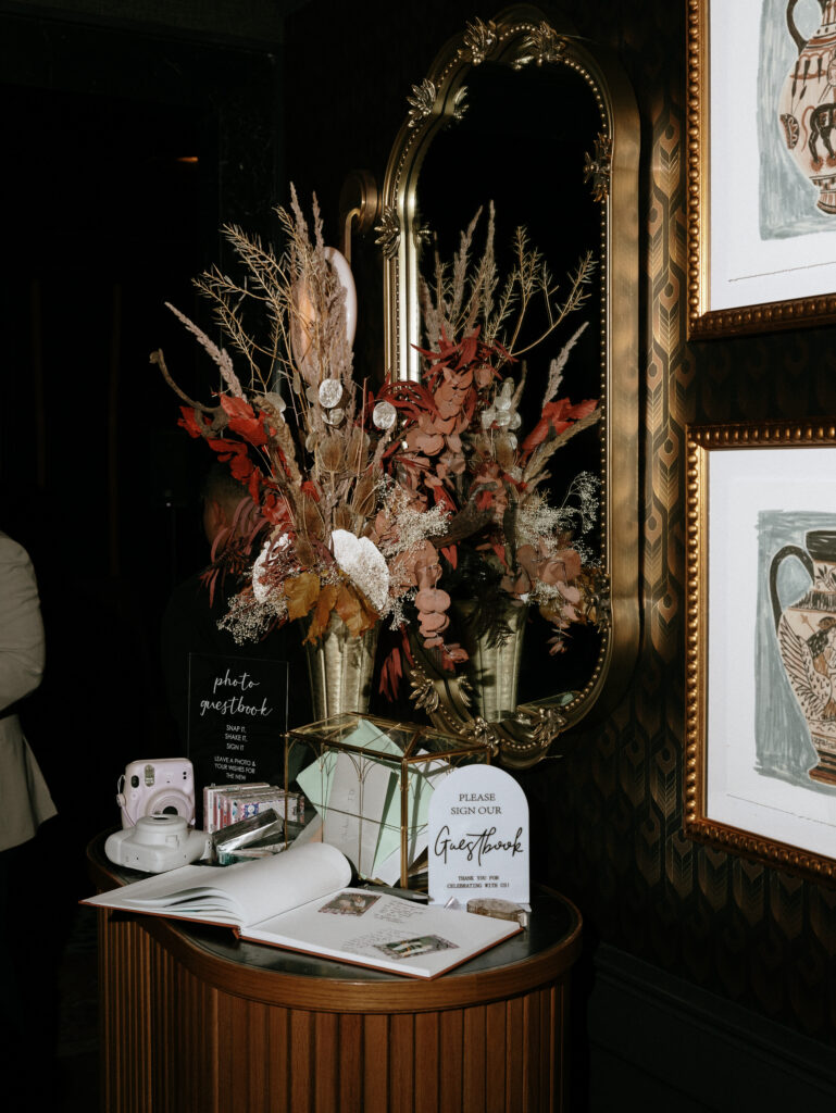 Ornate gold oval mirror with a dried flower arrangement in front, set against a dark patterned wall and a guestbook table nearby.