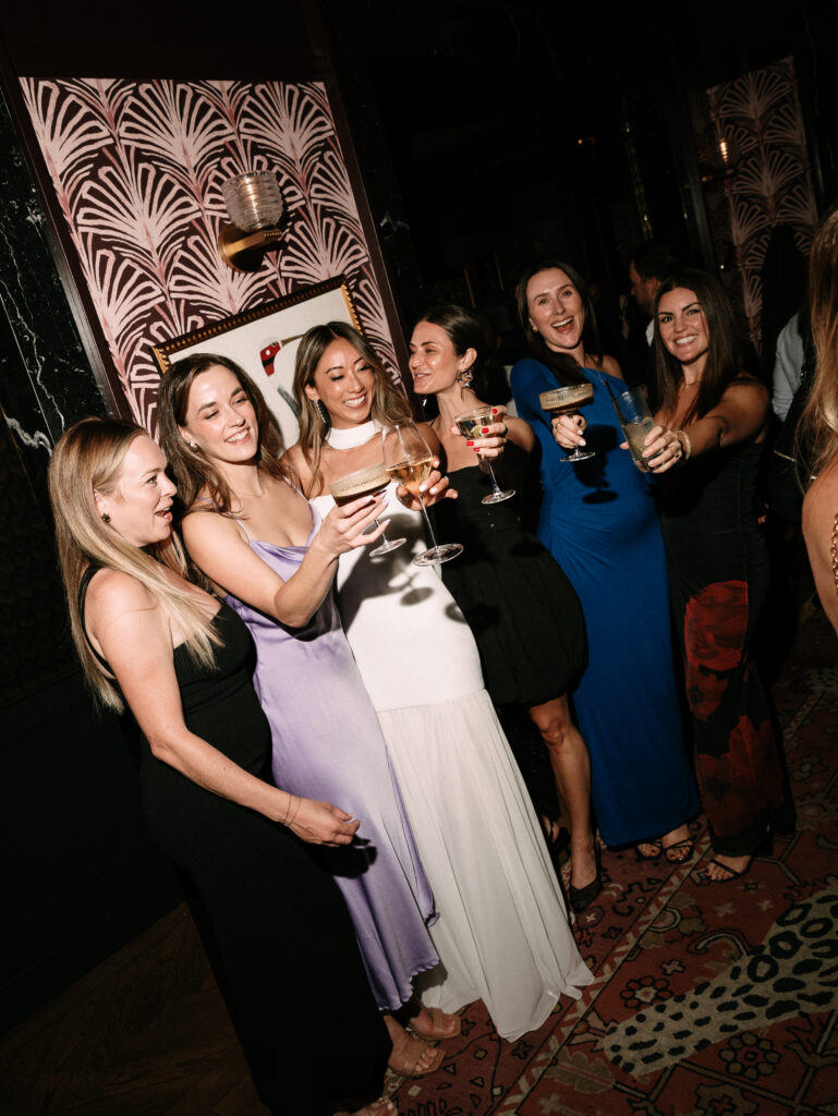 Group of women in evening dresses toasting with cocktails at a party. Pipelines, smiles, and raised glasses against patterned wall background.