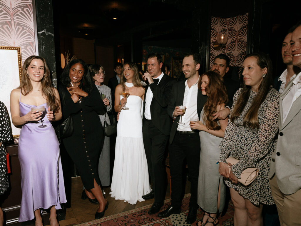 Group of well-dressed friends at a party posing for a photo, holding drinks and smiling indoors.