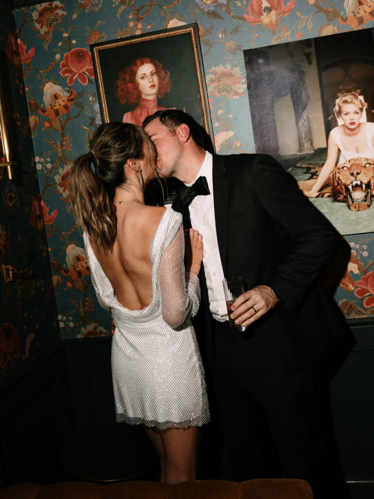 Couple in formal attire kissing against a wallpapered wall with framed portraits in the background, one holding a drink.
