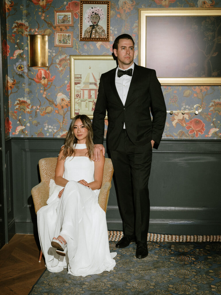 Man in a black tuxedo stands beside a seated woman in a white gown in a vintage, floral-patterned room.