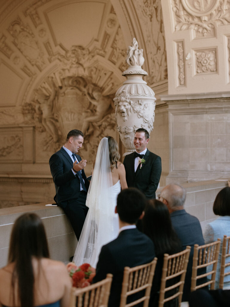 Bride and groom exchange vows in a grand, ornate hall with a stone urn centerpiece behind them.