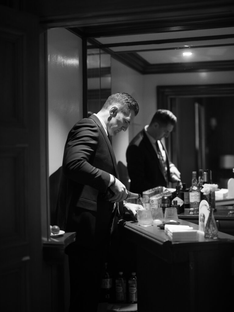 Black-and-white photo of a man in a suit pouring a drink at a bar, with bottles and glasses on the counter nearby.