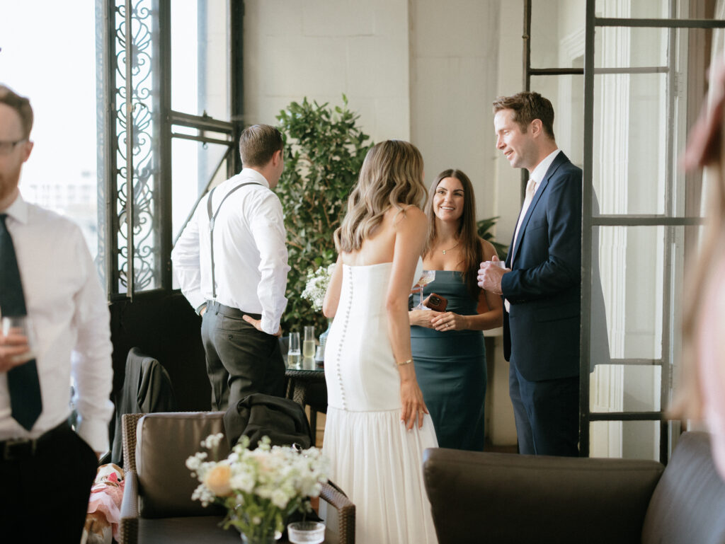 Group of formally dressed guests chatting and holding drinks at a bright indoor reception with plants in the background.