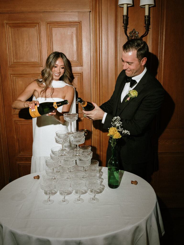 Bride and groom pour champagne into a crystal glasses tower at a reception, smiling in an elegant wood-paneled room.
