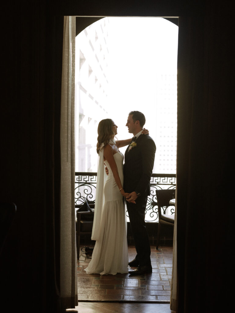 Bride and groom embrace and smile at each other in a doorway, backlit by bright exterior light behind them.