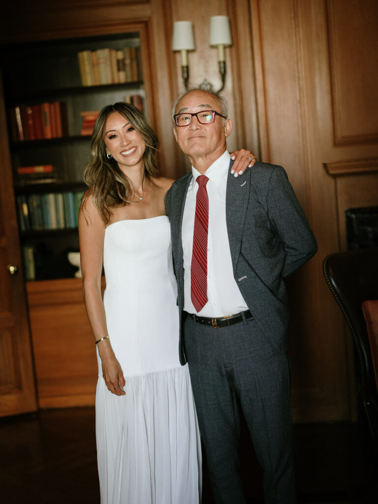 Smiling couple posing in a wood-paneled room; woman in a white strapless dress and man in a gray suit with a red tie, with bookshelves in the background