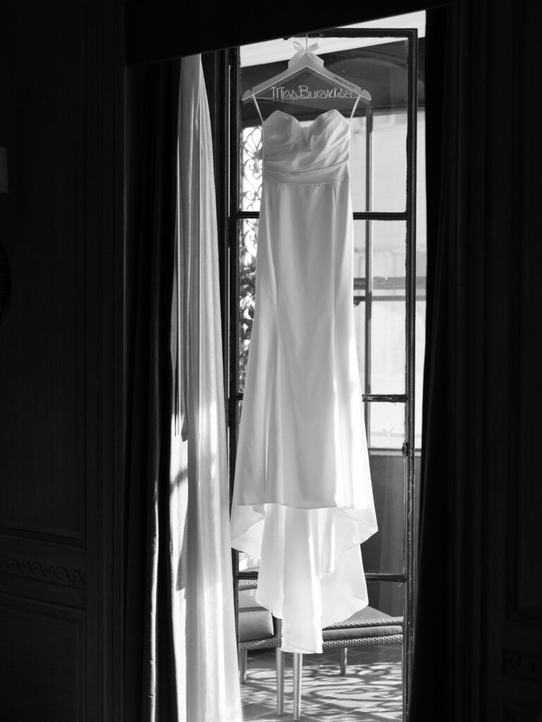 Wedding gown on a hanger hanging in front of a sunlit window, viewed through an open doorway (black and white).