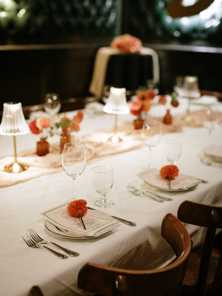 Elegant dining table with white linens, orange flowers, and neatly arranged place settings at dusk.