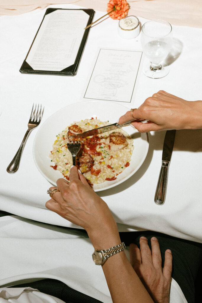 Person cutting a plate of risotto with meat pieces at an elegant table setting, with a menu, glass, and a pink flower nearby.