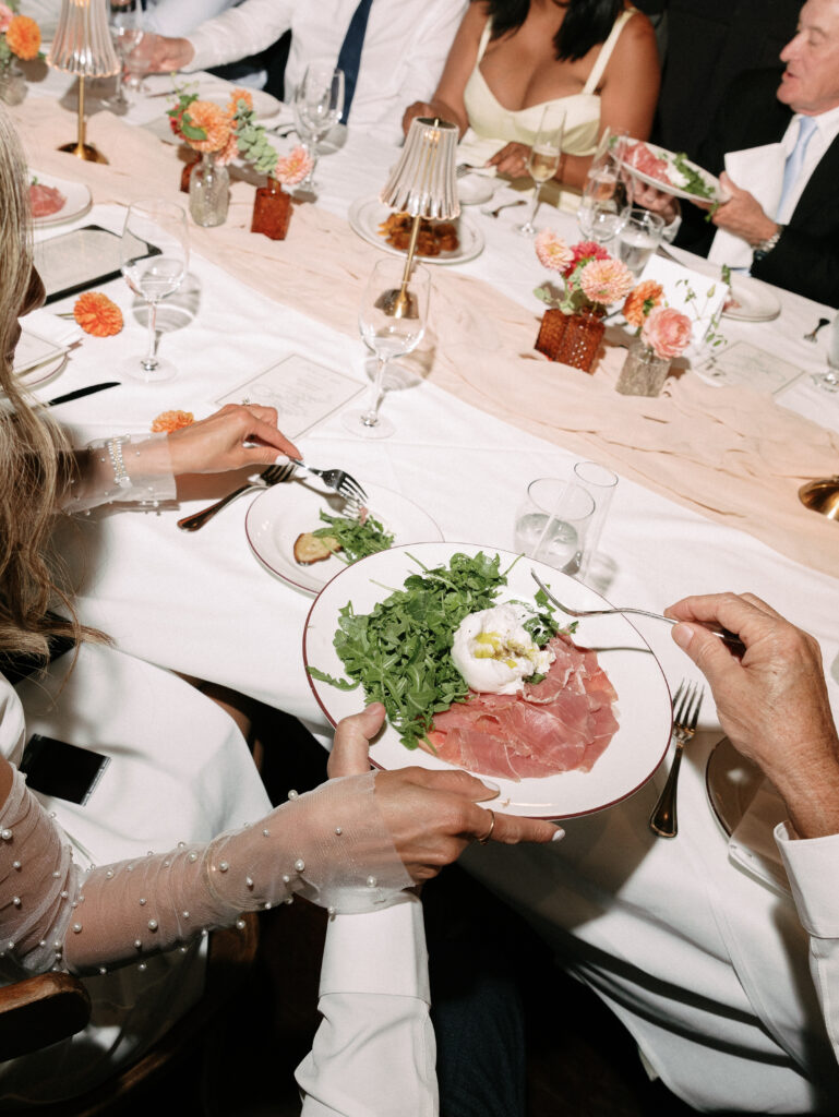 Guests at a formal dinner share a plate of greens, prosciutto, and a poached egg, with others eating in the background.
