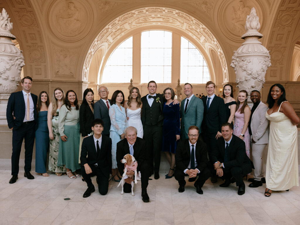 Group of formally dressed people posing for a photo on marble steps inside an ornate arched hall, with a dog at the front wearing a pink collar.