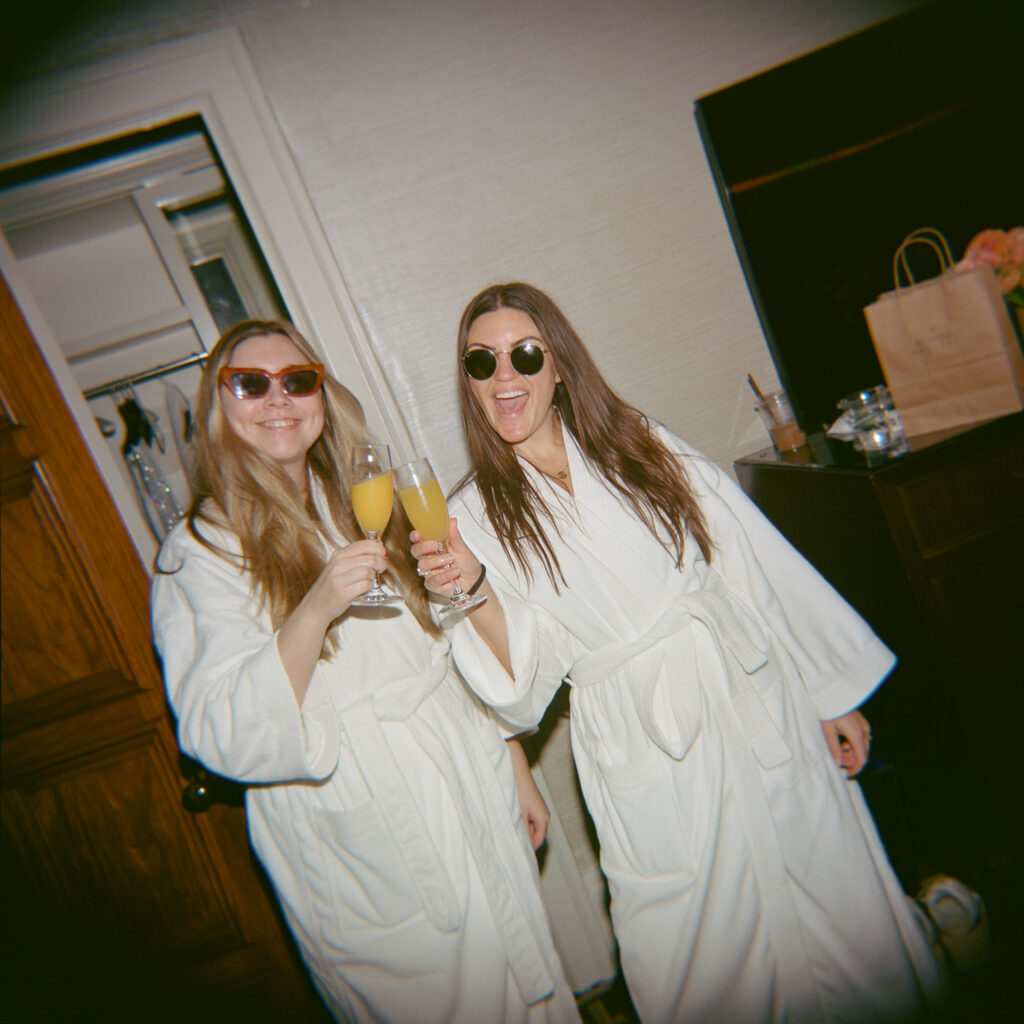 Two women in white robes toast with champagne glasses indoors, wearing sunglasses.