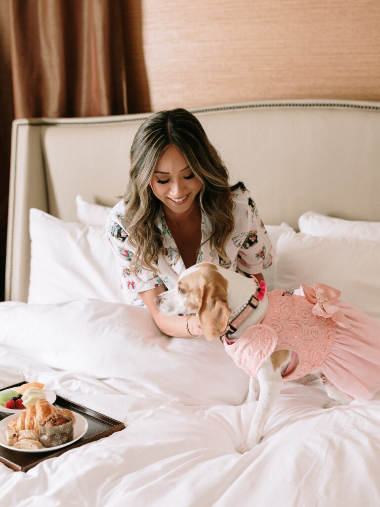 Woman smiles at a small dog in a pink dress on a bed with a tray of pastries nearby.