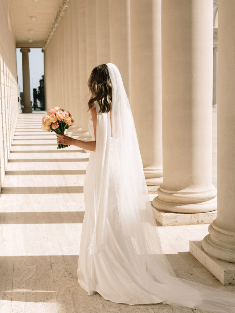 Bride in a white gown with veil, holding a pastel bouquet, stands beside tall beige columns in a sunlit colonnade.