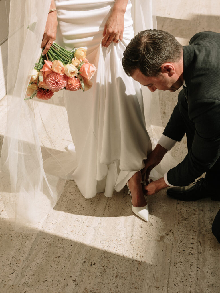 Bride in a white dress with veil, holding a pink-peach bouquet while a man in a dark suit adjusts her white heel.