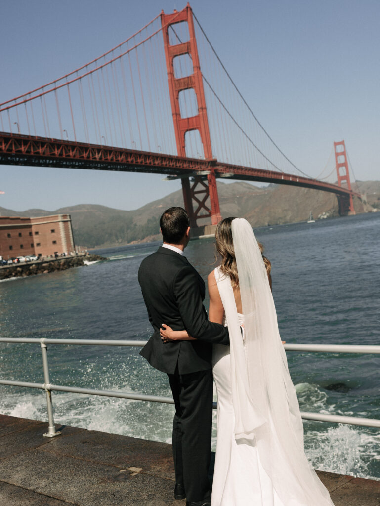 Bride and groom in wedding attire stand with backs to the camera, overlooking the water with the Golden Gate Bridge in the background.