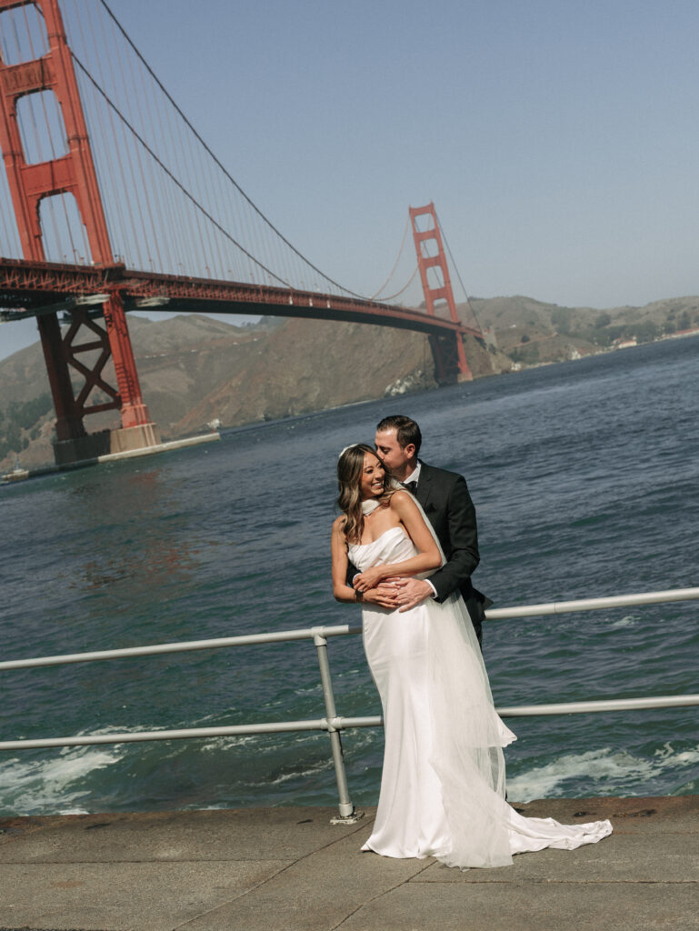 Bride in a white wedding dress and groom in a black tuxedo embrace at a railing by the water, Golden Gate Bridge in the background