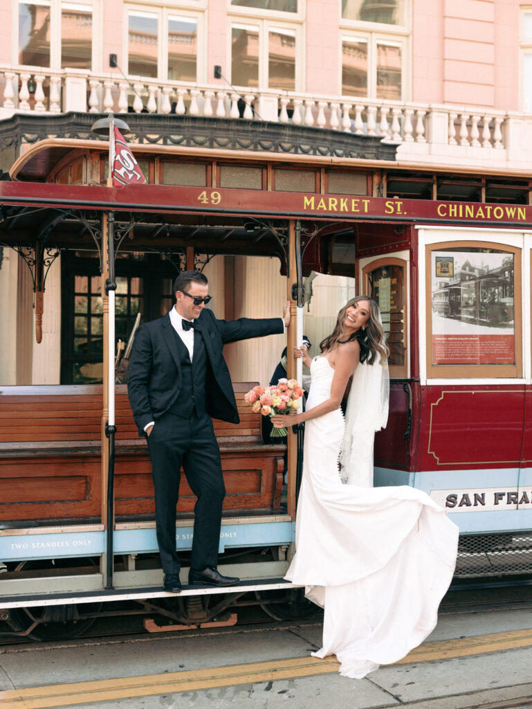 Bride in a white wedding gown and bouquet stands with groom in a tuxedo on a vintage trolley car labeled Market St. Chinatown, smiling at camera.