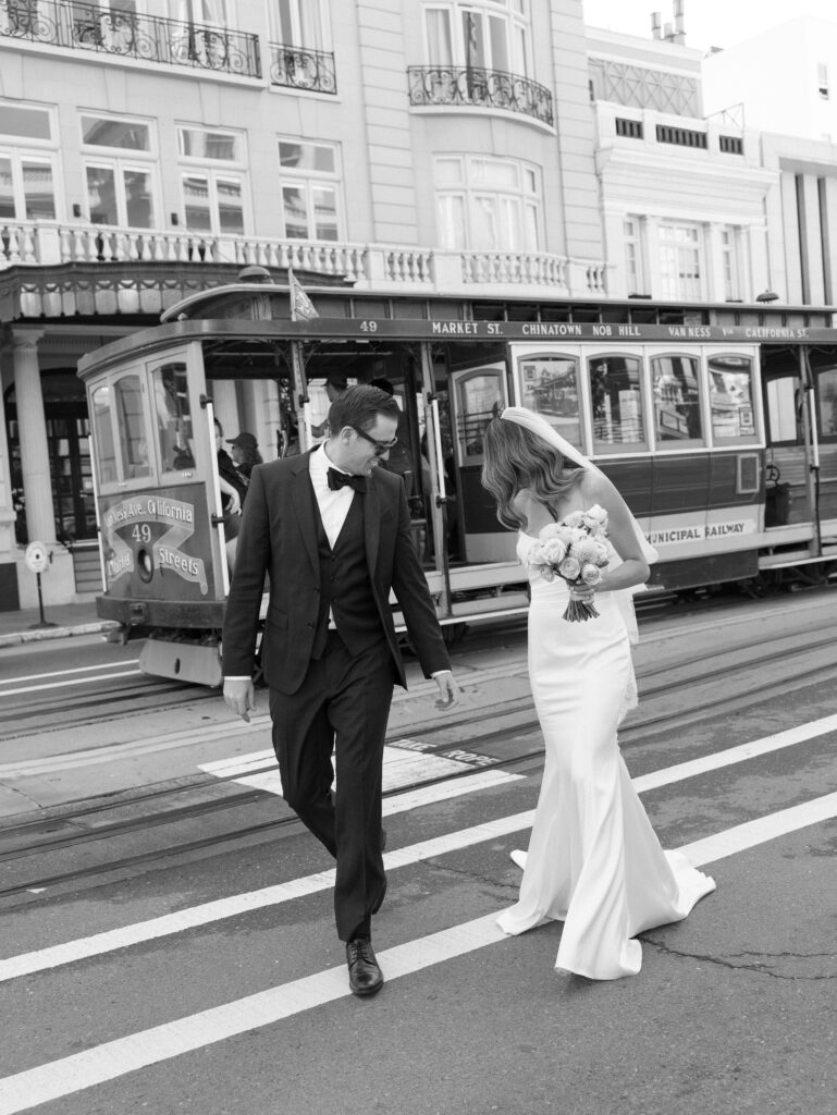 Bride in a white wedding dress with bouquet and a groom in a black suit walk past a vintage streetcar on a city street.