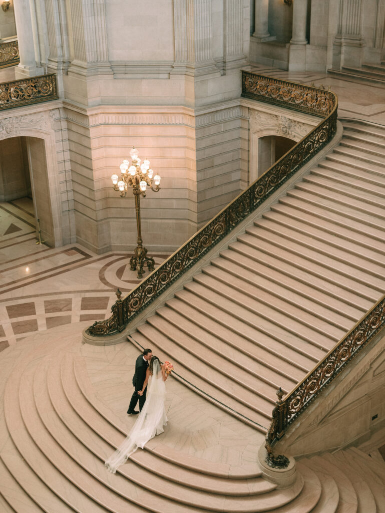A bride and groom stand at the bottom of a grand marble staircase under a chandelier in a stately, classical interior 1st floor hall.