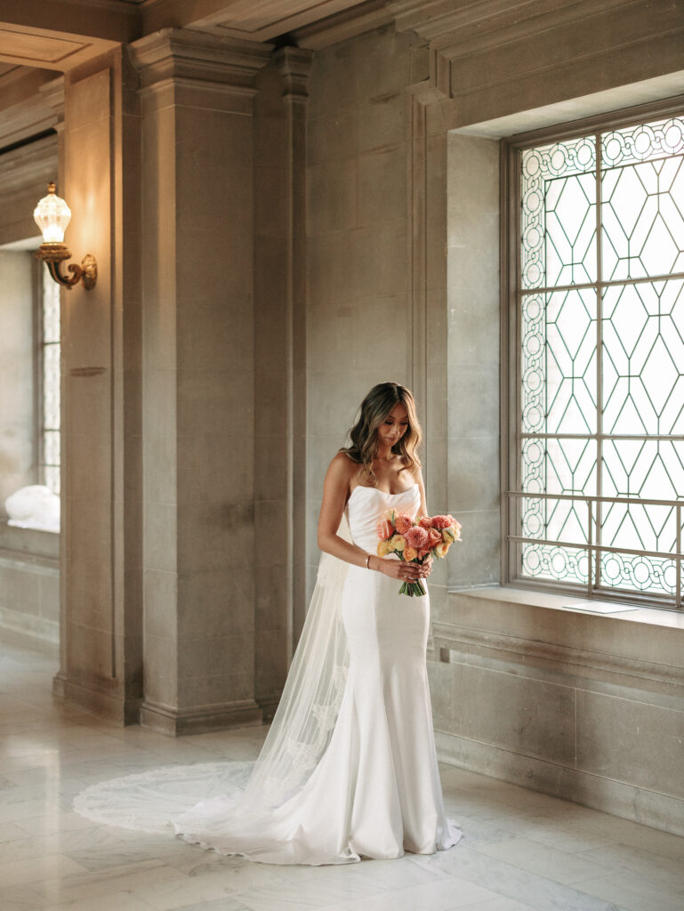 Bride in a white wedding gown with a veil holds a pink-peach bouquet in a stone hallway beside a large decorative window.