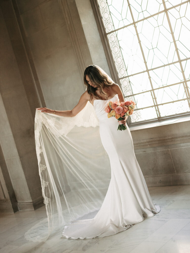 Bride in a white wedding gown holding a pastel bouquet, lace veil trailing as she stands in a sunlit marble hall near a decorative window.
