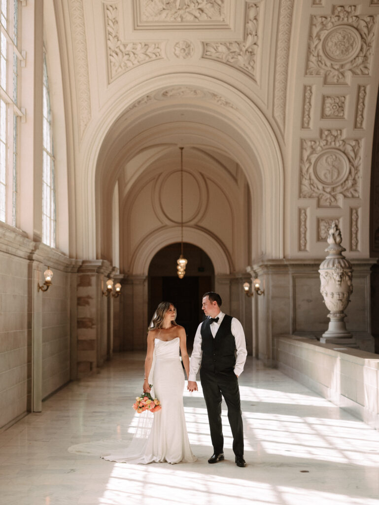 Bride in a white gown and groom in a black vest walk hand in hand through a sunlit, ornate marble hallway with arches and decorative walls.