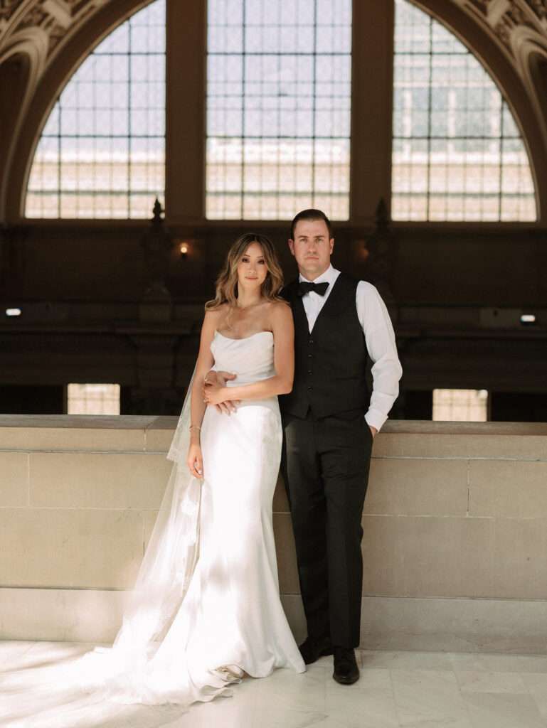 Bride in a white wedding gown with a veil and a groom in a black vest and bow tie stand together on marble steps in a grand arched hall behind them.",