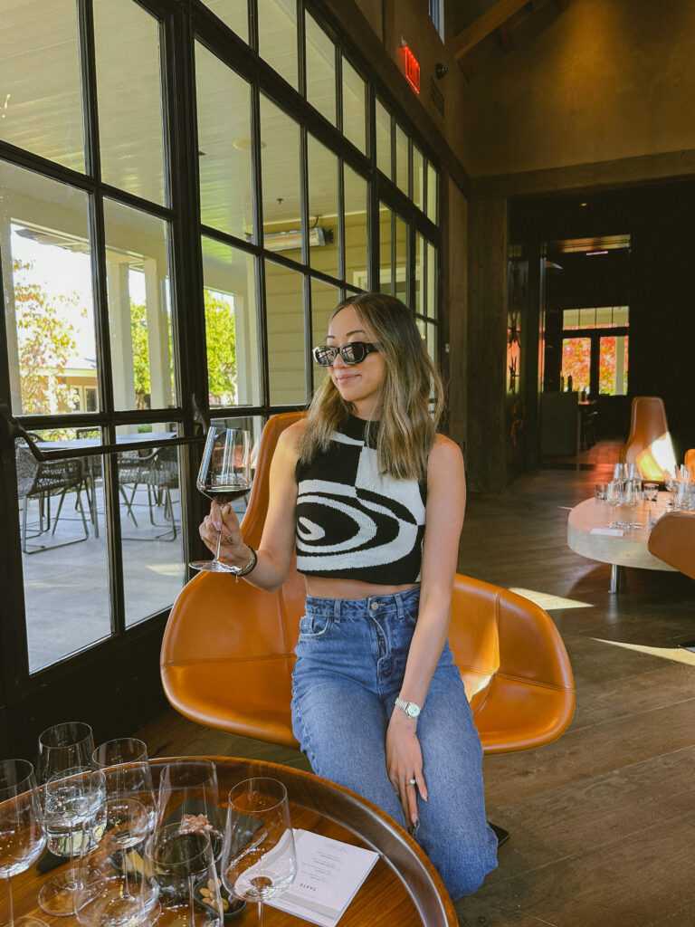 Woman wearing sunglasses and a black-and-white patterned sleeveless top sits in an orange chair at a dining table, holding a glass of red wine.