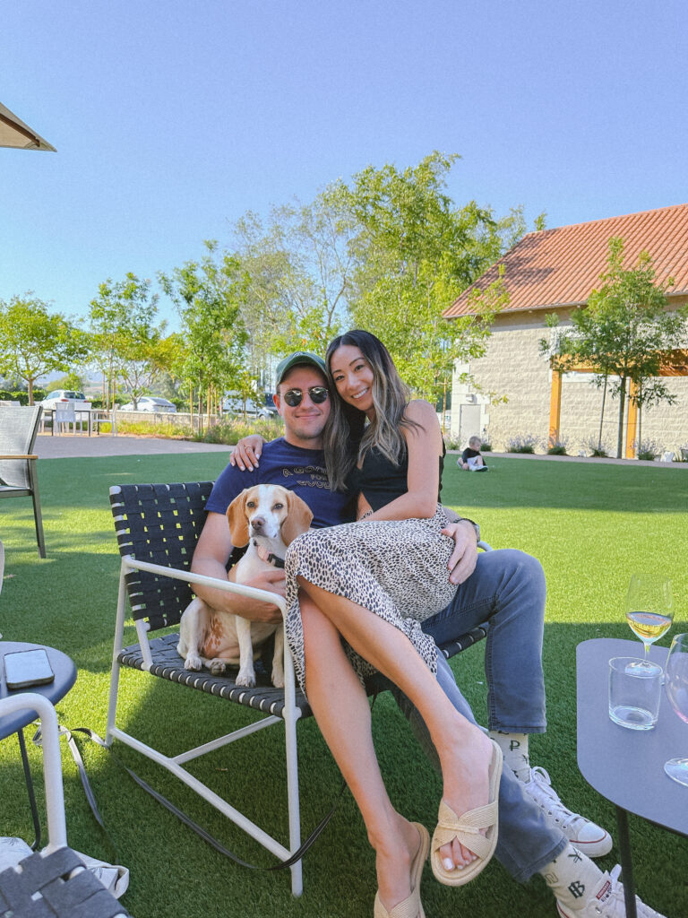 Couple sitting on a patio chair outdoors with a beagle on their laps, smiling on a sunny day with green grass and trees behind them.