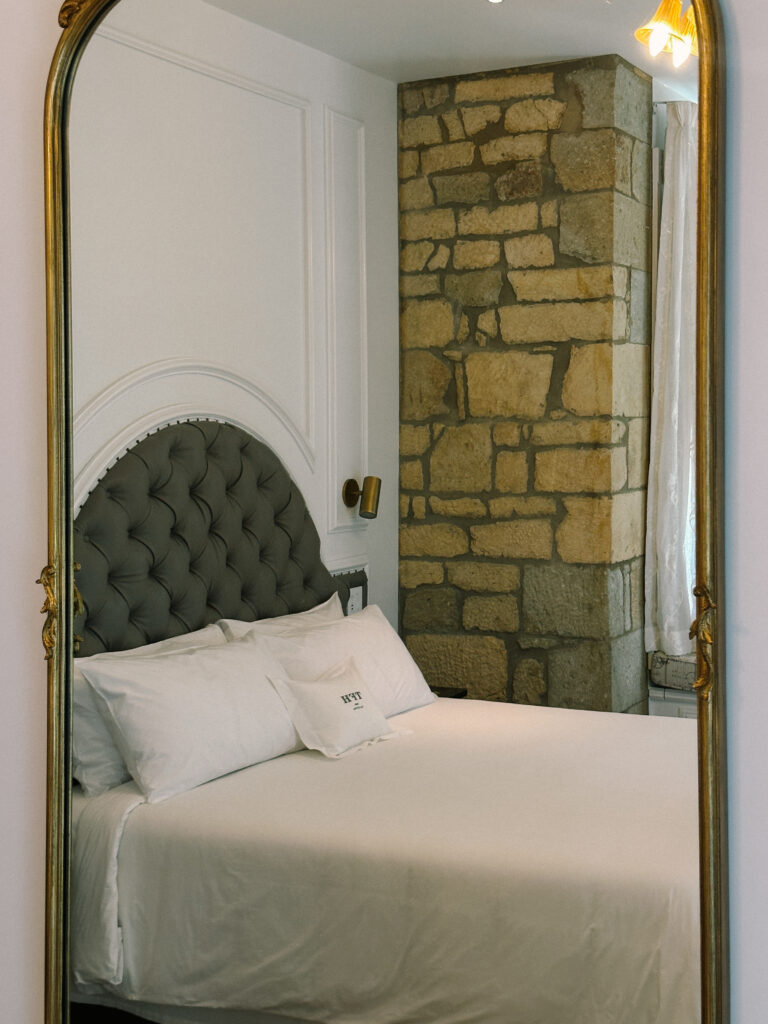 Bedroom corner with tufted gray headboard, white bedding, a stone brick pillar, and a gold-framed mirror