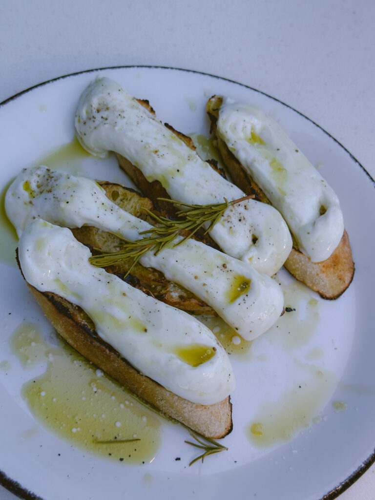 Toasted bread slices topped with poached eggs, rosemary sprig, and olive oil on a white plate.