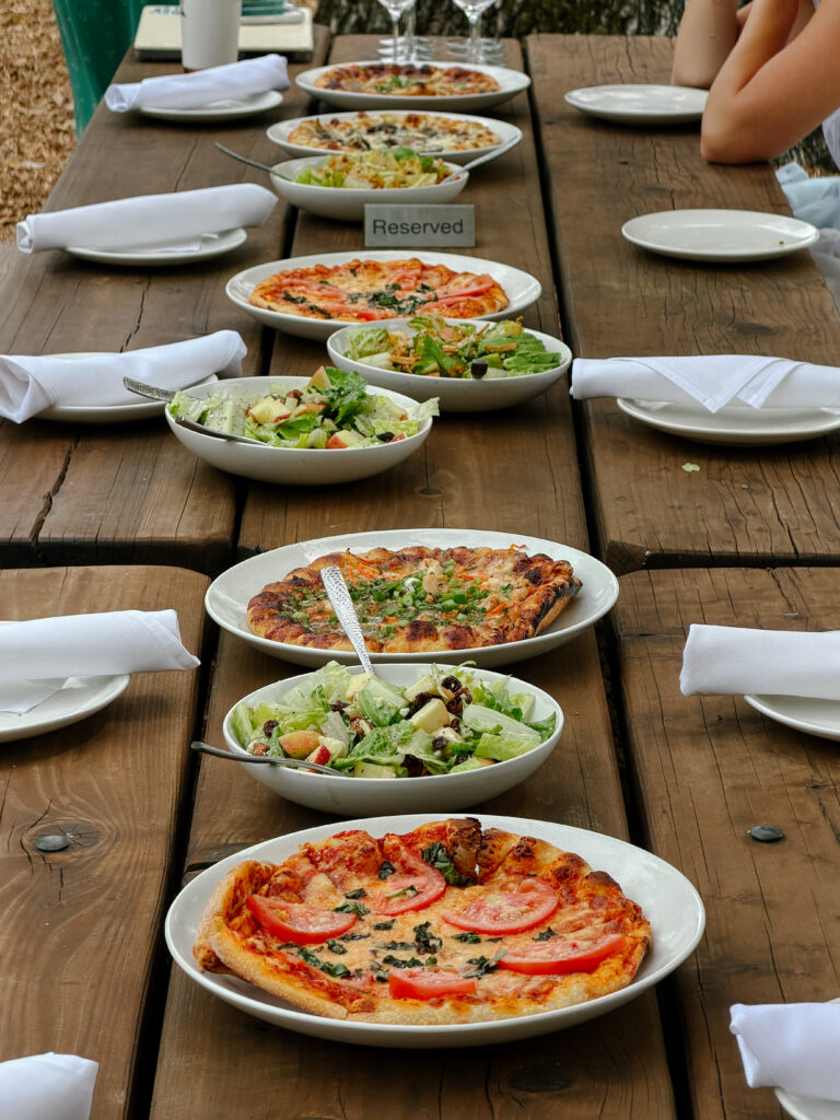 Outdoor meal setup: pizzas and bowls of salad arranged along a long wooden picnic table, with a 'Reserved' sign in the center.