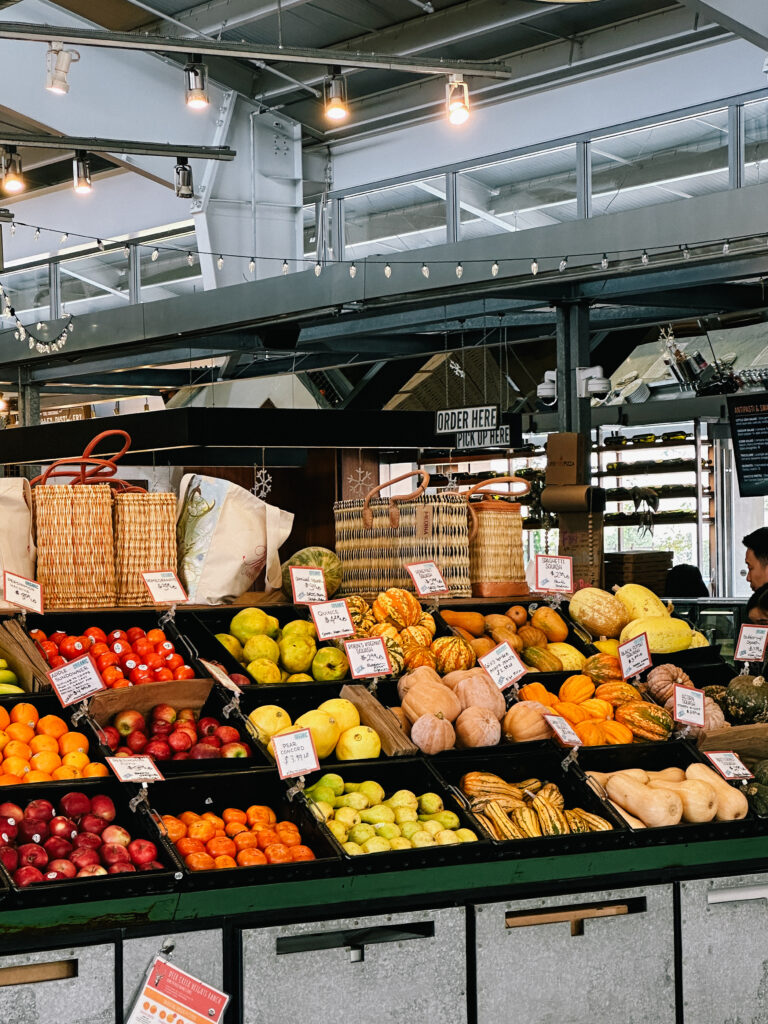 Indoor market fruit stand with apples, oranges, pears, and gourds on display under string lights.
