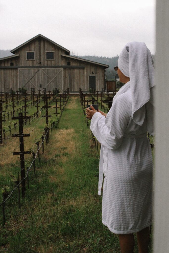 Person in a white bathrobe with a towel on their head stands beside a graveyard of wooden crosses, facing a weathered wooden barn in a rural setting.