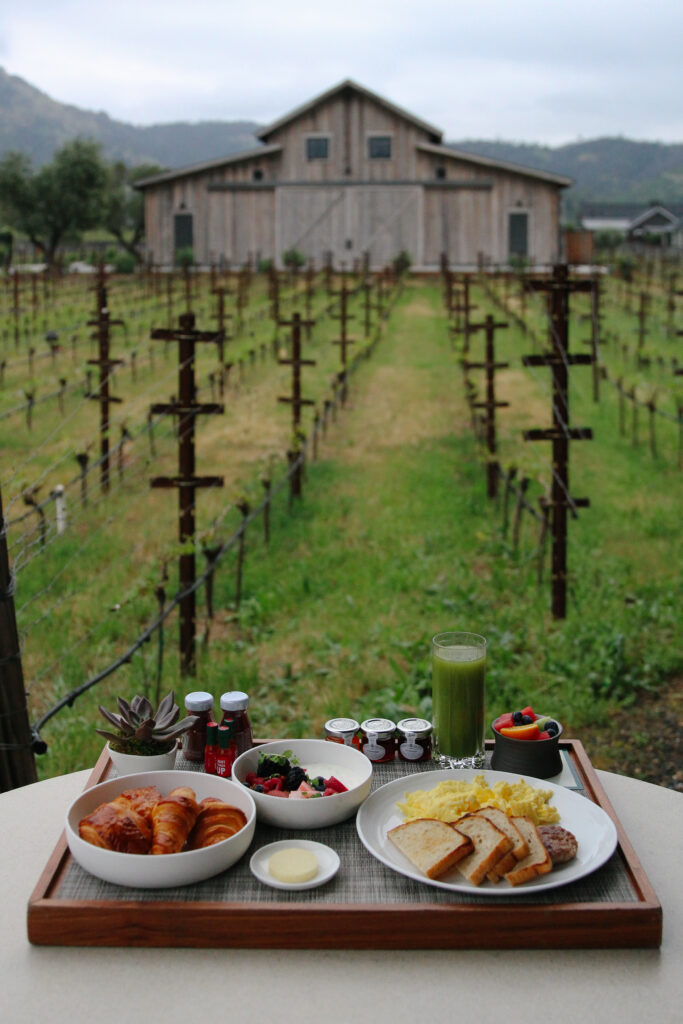 Outdoor breakfast spread on a wooden tray with croissants, fruit, jam, and green smoothie, set before a rustic barn and vineyard fencing.