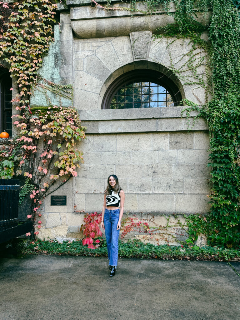 A young woman stands in front of a stone wall with a large arched window, ivy climbing the walls and autumn leaves nearby.**