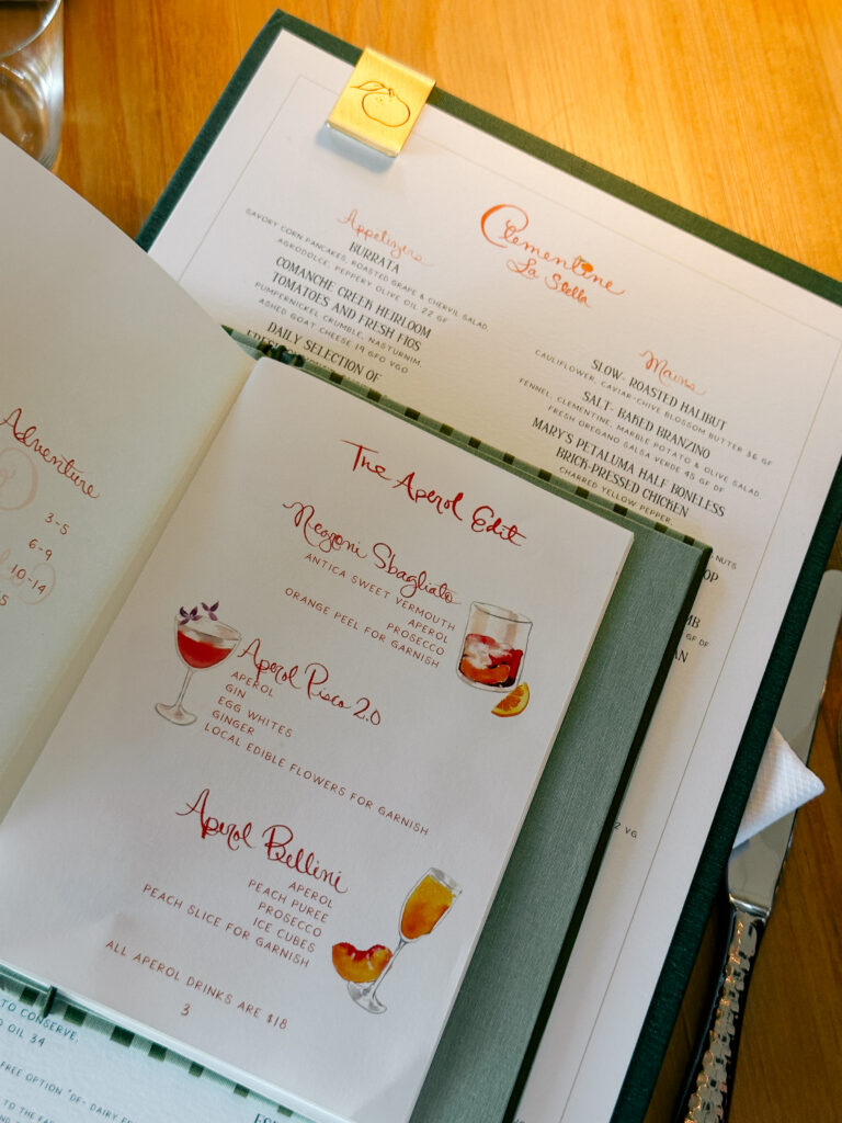 Open menu laid on a green binder with red handwritten headings and small cocktail illustrations on white pages.
