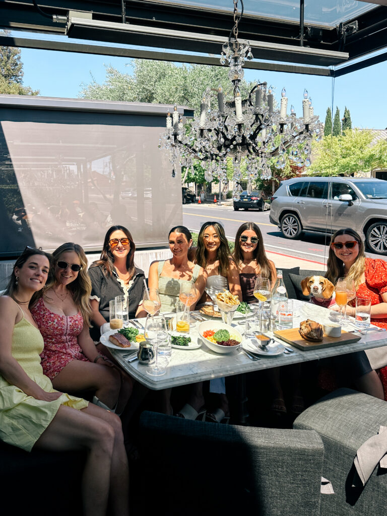 Group of friends dining at an outdoor table under a chandelier, plates of food and a small dog nearby on a sunny street.