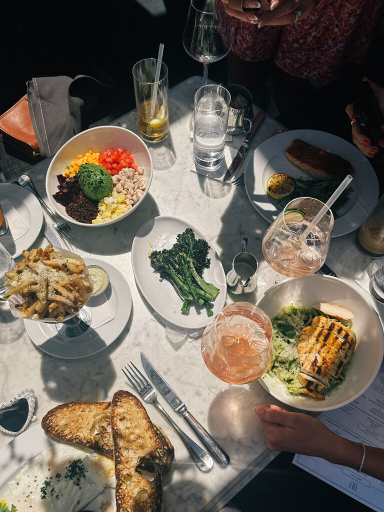 Overhead view of a marble table set for a meal: a colorful salad bowl with avocado, greens, bread, and drinks nearby.