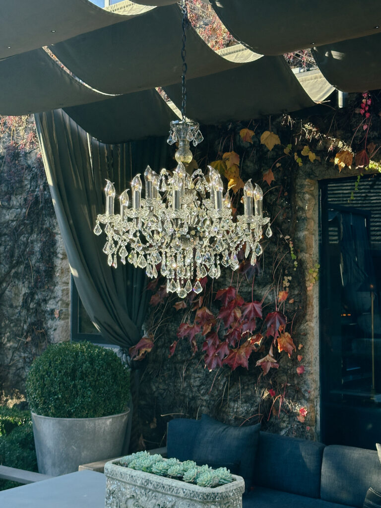 Crystal chandelier hanging outdoors above a grey couch in a patio area with ivy-covered stone wall.