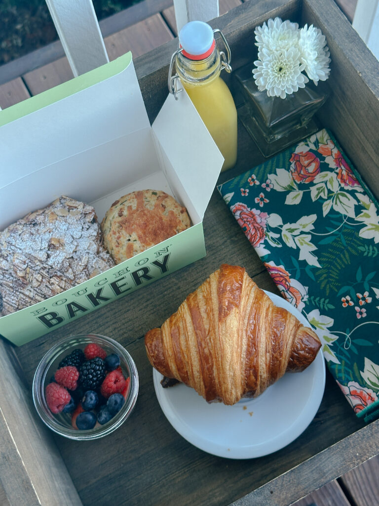 Brunch spread with a croissant on a white plate, pastries in a green bakery box, a cup of mixed berries, and a bottle of orange juice nearby in a wooden tray with a vase of white flowers and a floral napkin.