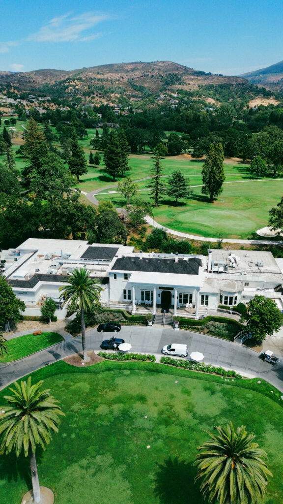 Aerial view of a white, mansion-like building next to a bright green golf course with scattered palm trees and distant hills under a blue sky.