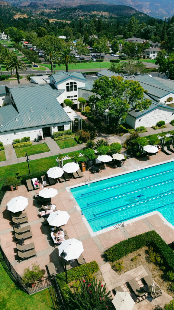 Aerial view of a resort pool deck with loungers and white umbrellas, surrounded by white buildings, palm trees, and landscaped gardens.