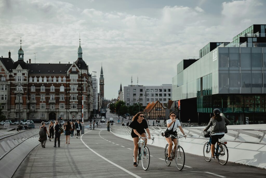 People cycling and walking on a wide city bridge; left is historic brick buildings, right a modern glass building against a blue sky with clouds.