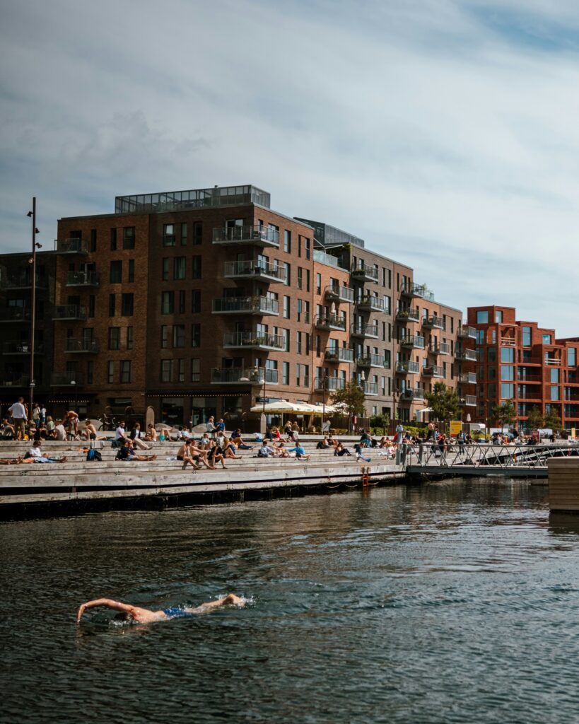 People relaxing on concrete steps by the water, with a brick apartment building and balconies behind them; a swimmer is in the foreground of the river/harbor.