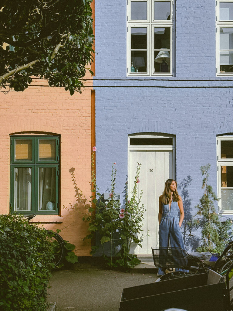 Woman in a blue jumpsuit stands outside a two-tone peach-and-lavender brick house, with white-framed windows and greenery nearby.